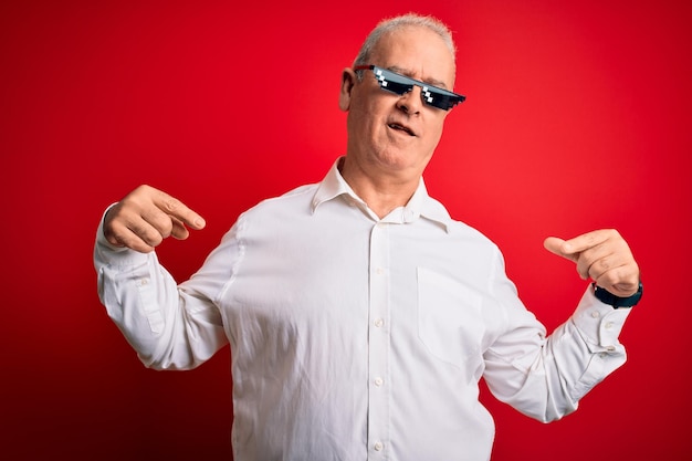 Sędziwy mężczyzna w średnim wieku w śmiesznych okularach przeciwsłonecznych na odizolowanym czerwonym tle, wyglądający pewnie z uśmiechem na twarzy, wskazujący na siebie palcami dumnymi i szczęśliwymi