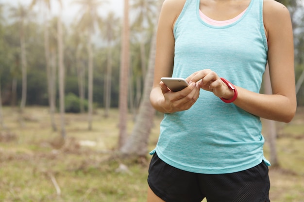 Sekcja środkowa biegaczki w stroju sportowym trzymająca telefon komórkowy, korzystająca z aplikacji do monitorowania kondycji do monitorowania postępów odchudzania podczas treningu cardio.