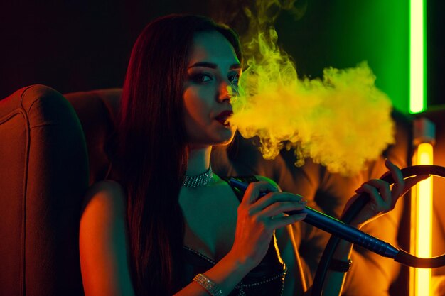 Seksowna brunetka modelka pali fajkę wodną, wydychając dym w luksusowym klubie nocnym.