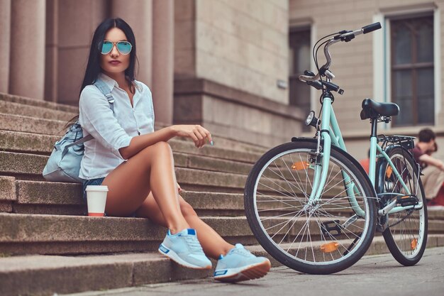 Seksowna brunetka ubrana w bluzkę i dżinsowe szorty w okularach przeciwsłonecznych, relaksująca się po jeździe na rowerze, siedząca przy filiżance kawy na schodach w mieście.
