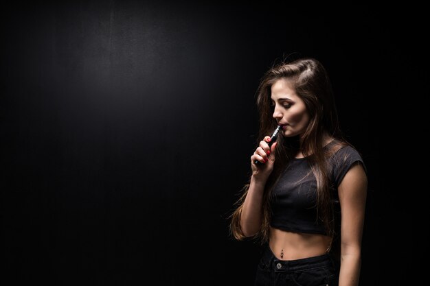 Seksowna dziewczyna w czarnej sukni palenia papierosów elektronicznych na ciemnej ścianie