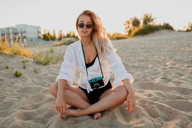 Seksowna Podróżująca Kobieta Relaks Na Plaży W Ciepły Letni Wieczór. Siedząc Na Piasku. Ubrana W Białą Bluzkę I Okulary Przeciwsłoneczne. Trzymając Aparat Retro.