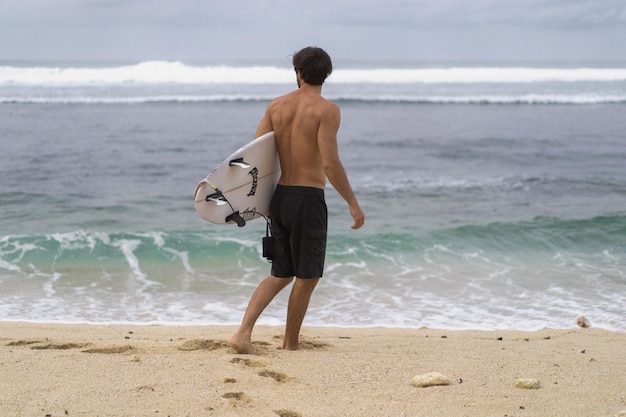 Seksowny surfer surfing człowiek z deską surfingową. Przystojny młody sportowiec mężczyzna trzyma deskę surfingową z mokrymi włosami na wakacje sport lato plaża. Sportowy cel podróży. Styl życia surfingu.