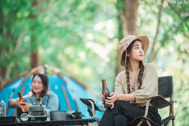 Selektywny fokus Młoda Azjatycka ładna kobieta i jej przyjaciółki podróżujące odpoczywają na kempingowych krzesłach w namiocie Dopingują i piją piwo podczas biwakowania rozmawiając z zabawą i radością