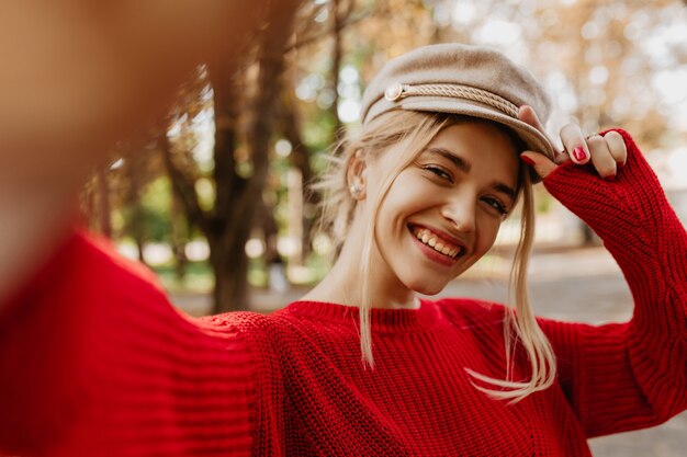 Selfie z piękną blondynką uśmiechniętą w parku. Oszałamiająca kobieta w sezonowych ubraniach, zabawy jesienią.