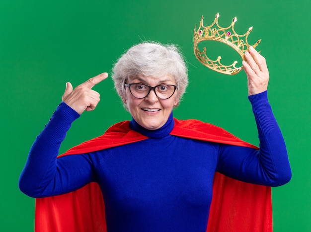 Senior kobieta superbohater ubrany w czerwoną pelerynę i okulary trzymające koronę wskazującą palcem wskazującym na głowę uśmiechający się pewnie w dół stojący na zielonym tle