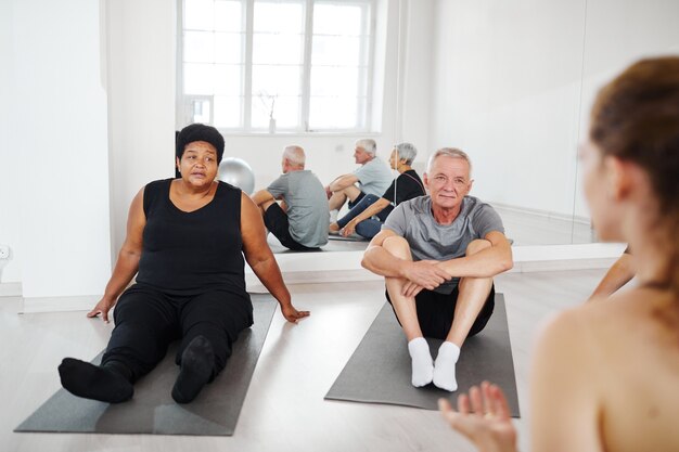 Seniorzy wykonujący ćwiczenia relaksacyjne na podłodze i słuchający trenera podczas gimnastyki...