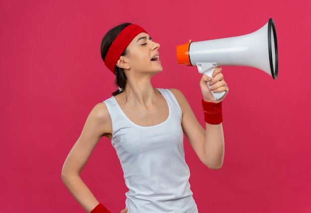 Sfrustrowana młoda dziewczyna fitness w odzieży sportowej z opaską krzyczy do megafonu stojącego nad różową ścianą