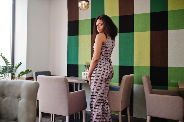 Sharming elegancka młoda afroamerykanka z długimi kręconymi włosami w kombinezonie pozującym w kawiarni w pomieszczeniu