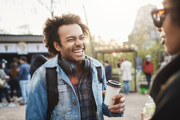 Sideview portret modnego słodkiego afrykańskiego faceta z afro fryzurą śmiejącego się głośno nad żartem podczas rozmowy z przyjacielem w parku, pijąc kawę i będąc optymistycznym Koncepcja przyjaźni