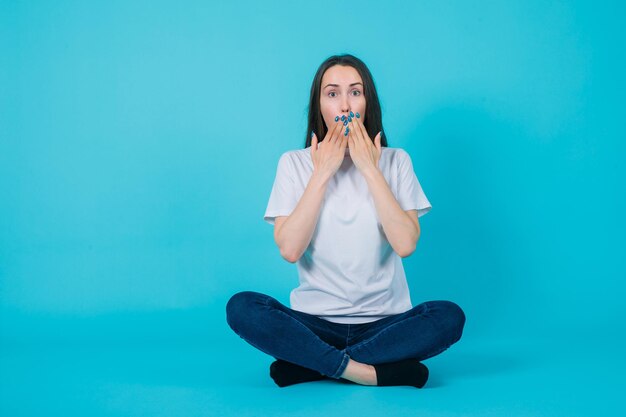 Siedząca na podłodze dziewczyna trzyma ręce na ustach na niebieskim tle