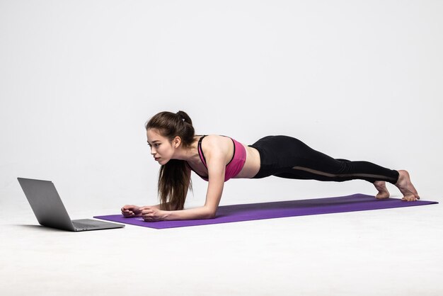 Silna wysportowana kobieta ćwiczy na macie do jogi przed laptopem, ubrana w sportowy strój na białym tle.