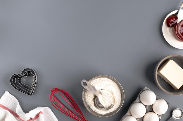 Składniki do pieczenia na ciemnoszarym tle: mąka, jajka, masło, kształt serca, trzepaczka, tekstylia. widok z góry. skopiuj miejsce. układ przepisu na ciasto lub ciasto z ciasteczkami