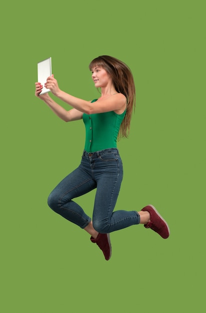 Skok młodej kobiety nad zielonym studio przy użyciu gadżetu laptopa lub tabletu podczas skakania.