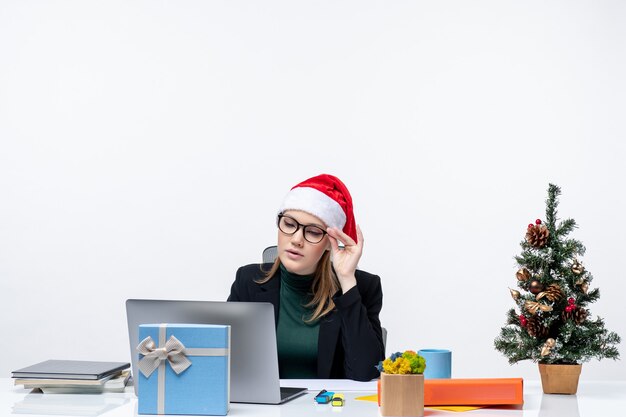 Skoncentrowana biznesowa kobieta z czapką Świętego Mikołaja siedząca przy stole z choinką i prezentem na niej i sprawdzająca jej pocztę na białym tle