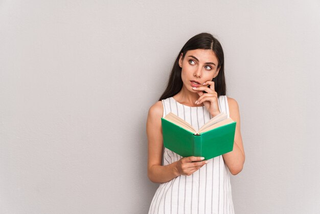 Skoncentrowana brunetka ubrana w sukienkę myśląca i patrząca na bok podczas czytania książki odizolowanej na szarej ścianie