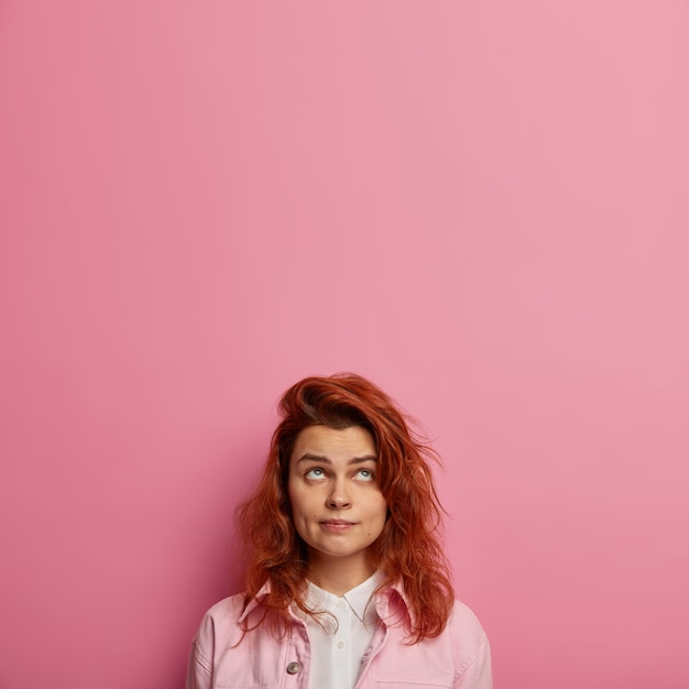 Bezpłatne zdjęcie skoncentrowana europejka o rudych włosach, zdrowej skórze, patrzy w górę, nosi różowe ubrania