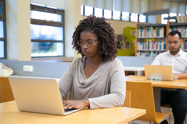 Skoncentrowana młoda kobieta pracuje z laptopem przy biblioteką