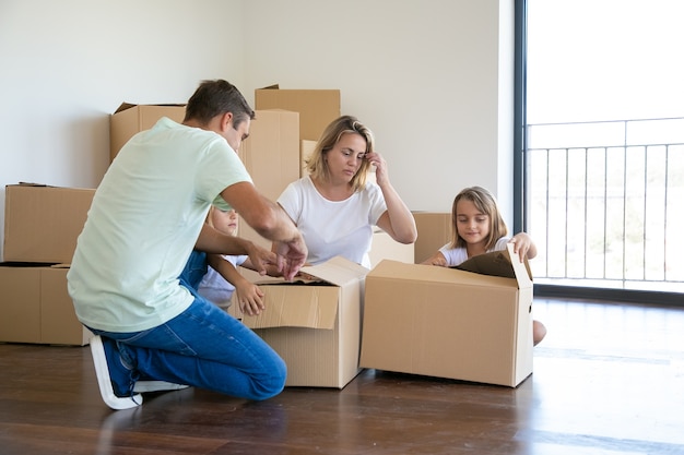 Skoncentrowani rodzice i dzieci rozpakowują rzeczy w nowym mieszkaniu, siadają na podłodze i otwierają pudełka