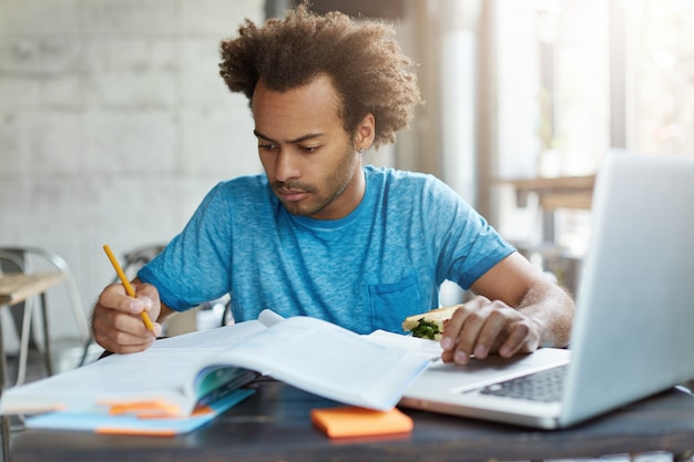 skoncentrowany Afroamerykanin hipster mężczyzna w niebieskiej koszulce przygotowuje się do egzaminu testowego zapisuje notatki w swoim zeszycie za pomocą laptopa do wyszukiwania potrzebnych informacji