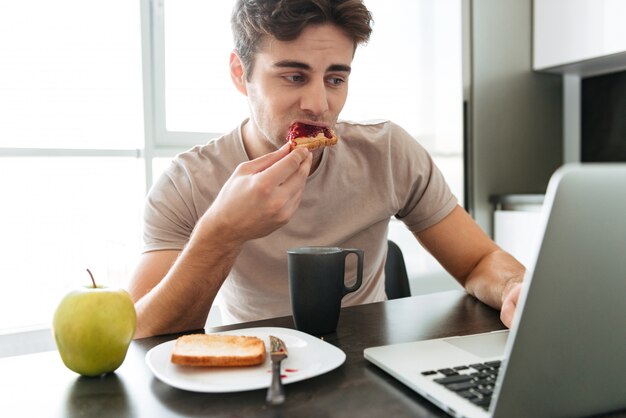 Skoncentrowany atrakcyjny mężczyzna używa laptop podczas gdy jedzący śniadanie