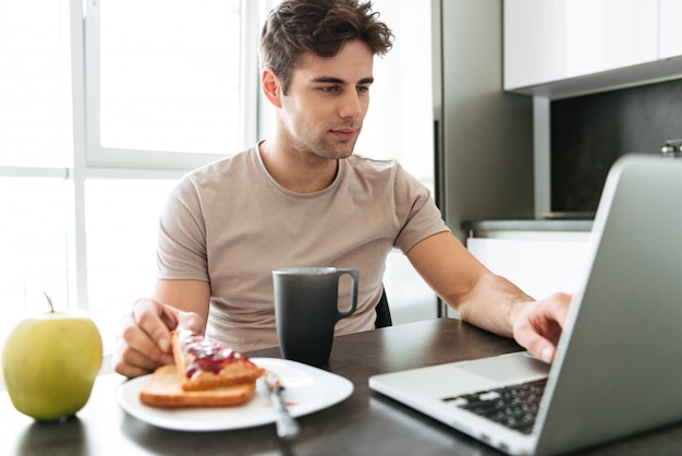 Skoncentrowany atrakcyjny mężczyzna używa laptop podczas gdy jedzący śniadanie
