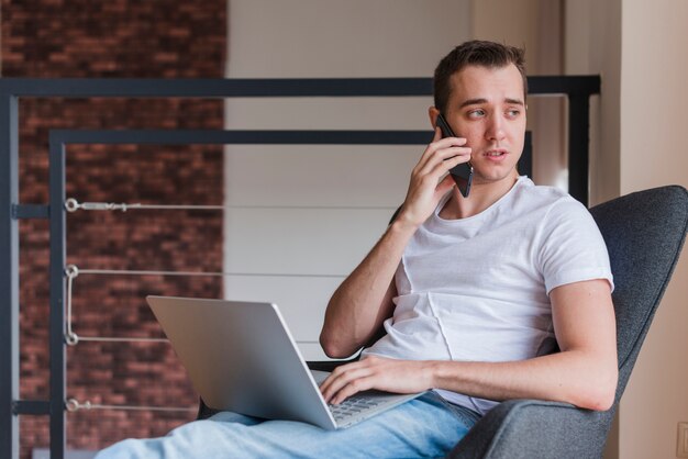 Skoncentrowany mężczyzna opowiada na telefonie komórkowym i obsiadaniu na krześle z laptopem