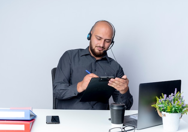 Skoncentrowany młody łysy mężczyzna call center noszenie zestawu słuchawkowego siedzi przy biurku z narzędzi pracy pisanie za pomocą pióra w schowku samodzielnie na białym tle