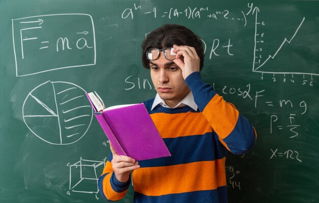 Skoncentrowany młody nauczyciel geometrii kaukaskiej w okularach, stojący przed tablicą w klasie, podnoszący okulary, czytający książkę