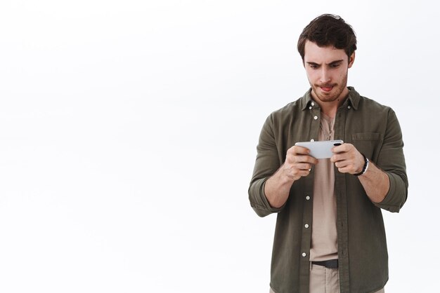 Skoncentrowany młody przystojny mężczyzna grający w intensywną grę trzymając telefon komórkowy poziomo lizać usta, pokazując język i marszcząc brwi, gdy wpatruje się w wyświetlacz smartfona, pobierz nowe zręcznościowe białe tło