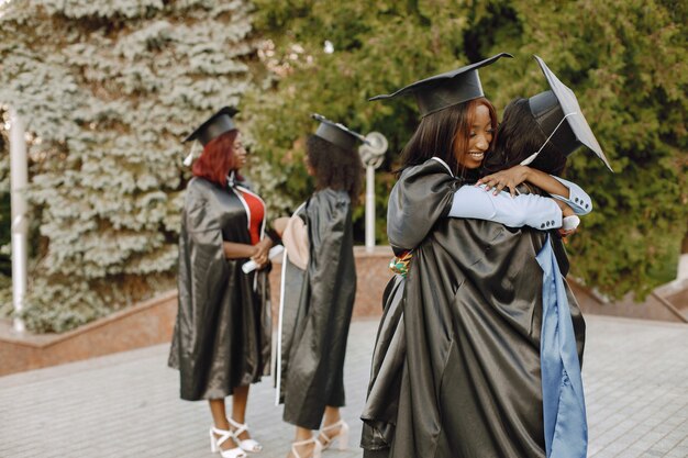 Skoncentruj się na dwóch młodych afro amerykańskich studentek ubranych w czarną suknię ukończenia szkoły. Kampus jako tło. Przytulające się dziewczyny