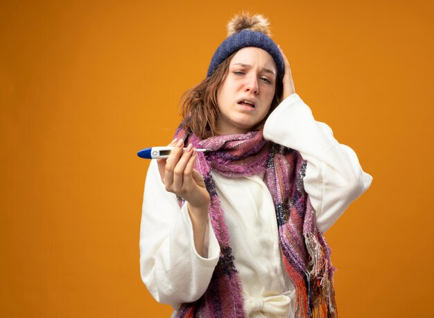 Słaba młoda chora dziewczyna w białej szacie i czapce zimowej z szalikiem trzymająca termometr kładąca rękę na głowie odizolowana na pomarańczowo z miejscem na kopię