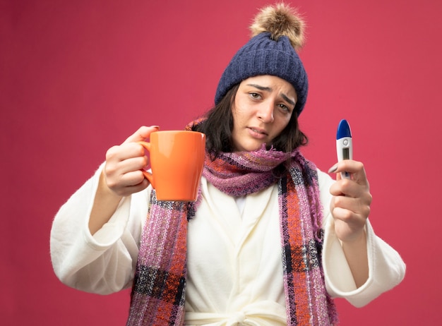 Bezpłatne zdjęcie słaba młoda kaukaska chora dziewczyna w czapce zimowej szaty i szaliku wyciągająca filiżankę herbaty i trzymająca termometr izolowany na szkarłatnej ścianie