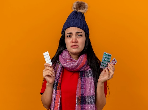 Słaba młoda kaukaski chora dziewczyna ubrana w czapkę zimową i szalik, trzymając paczki pigułek medycznych na białym tle na pomarańczowej ścianie z miejsca na kopię