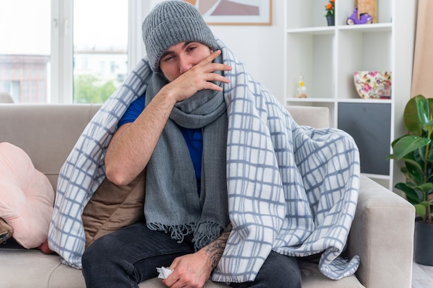 Słaby młody chory mężczyzna ubrany w szalik i czapkę zimową zawiniętą w koc, siedzący na kanapie w salonie, trzymający serwetkę i patrzący w kamerę, wycierając nos ręką z zamkniętym jednym okiem
