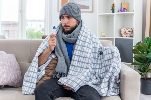 Słaby młody chory mężczyzna ubrany w szalik i czapkę zimową zawiniętą w koc, siedzący na kanapie w salonie, trzymający termometr patrząc w bok