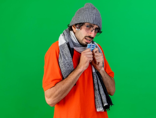 Bezpłatne zdjęcie słaby młody chory mężczyzna w okularach czapka zimowa i szalik trzymający paczkę kapsułek patrząc z przodu z paczką kapsułek pod kapeluszem odizolowanym na zielonej ścianie z miejscem na kopię