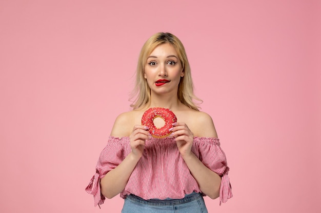 Śliczna blond laska z czerwoną szminką w różowej bluzce przygryza usta i trzyma pączka z jagodami