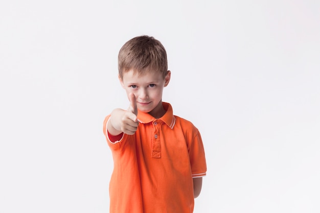 Bezpłatne zdjęcie Śliczna chłopiec jest ubranym pomarańczową koszulkę wskazuje przy kamerą na biel ścianie