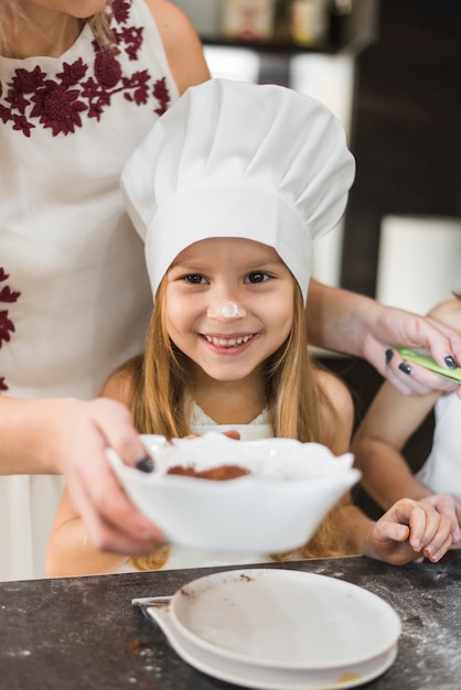 Śliczna dziewczyna jest ubranym szef kuchni kapeluszową pozycję w frontowej matce podczas gdy gotujący w kuchni
