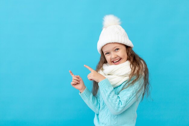 Śliczna dziewczynka dziecko w zimowe ubrania kapelusz i sweter wskazuje palce na coś na niebieskim tle na białym tle i uśmiecha się i śmieje się, miejsce lub miejsce na tekst