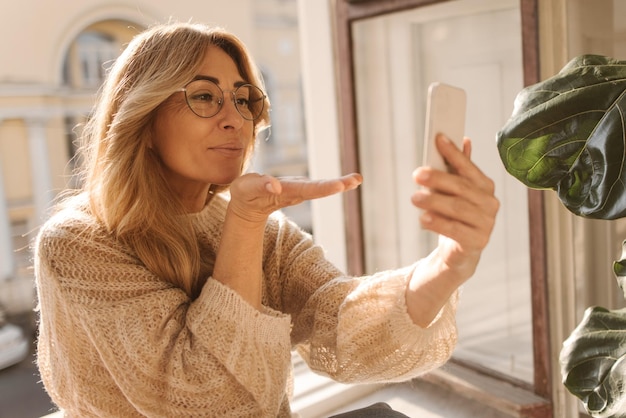 Śliczna kaukaska dorosła blondynka w ciepłym swetrze wysyła pocałunek powietrzny do telefonu trzymającego w dłoniach Koncepcja czasu selfie