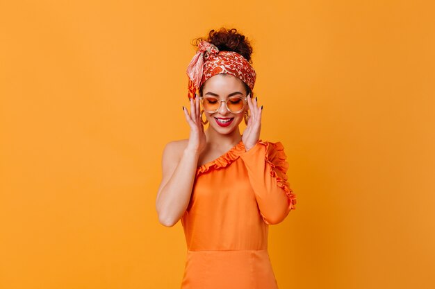 Śliczna kobieta w pomarańczowych okularach, jedwabnej sukience i opasce uśmiecha się na pomarańczowej przestrzeni.