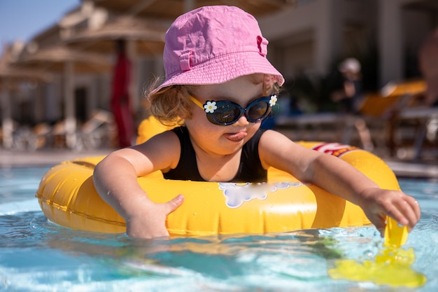 Śliczna mała dziewczynka w kapeluszu i okularach przeciwsłonecznych gra w basenie siedząc w kręgu pływania.