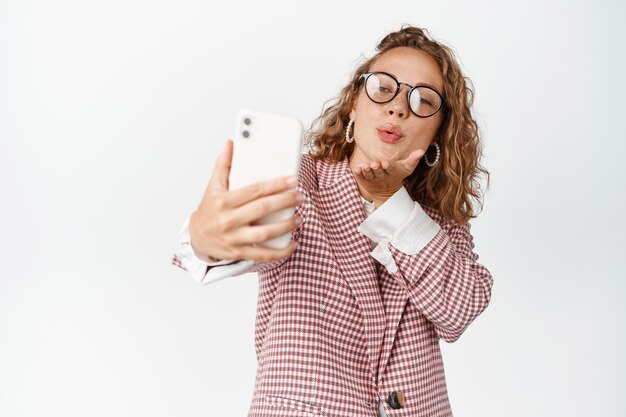 Śliczna menedżerka biorąca selfie, wysyłająca buziaki w aparacie telefonu komórkowego, romantyczne połączenie wideo w aplikacji smarpthone, stojąca na białym