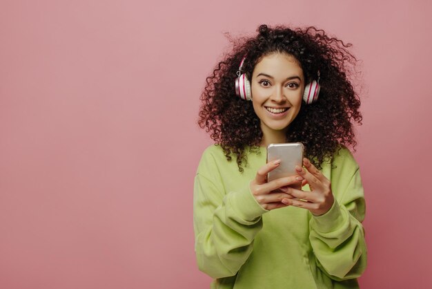Śliczna młoda brunetka kaukaska dziewczyna w słuchawkach patrzy na kamerę na różowym tle Kręcone piękno w zielonej kurtce trzyma telefon w dłoniach Koncepcja technologii i ludzi