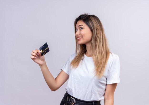 Śliczna młoda kobieta w białej koszulce uśmiecha się i pokazuje kartę kredytową na białej ścianie