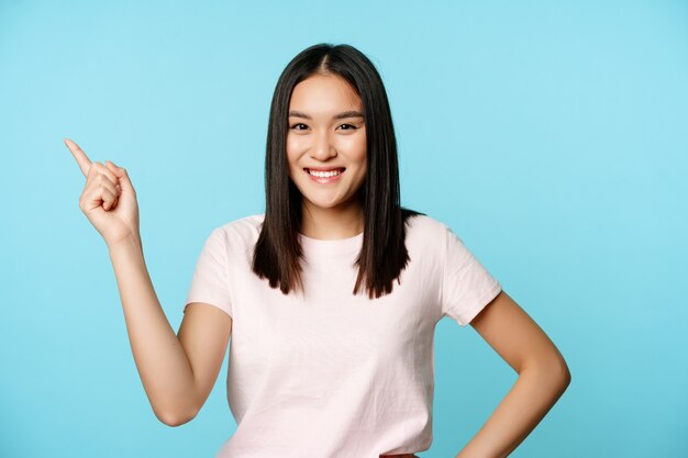 Śliczna uśmiechnięta studentka z azjatyckiej brunetki, wskazująca na lewy górny róg, pokazując baner promocyjny z nazwą firmy ...