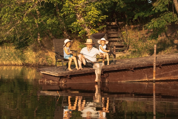 Śliczne dziewczynki i ich dziadek łowią ryby nad jeziorem lub rzeką. Odpoczynek na molo w pobliżu wody i lasu w porze zachodu słońca w letni dzień. Pojęcie rodziny, rekreacji, dzieciństwa, przyrody.