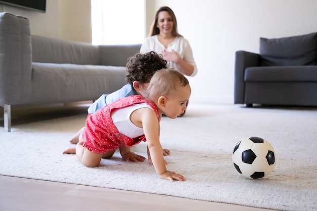 Bezpłatne zdjęcie Śliczne małe dzieci czołgają się na dywanie i bawią się piłką nożną. troskliwa matka siedzi na podłodze, uśmiechając się i obserwując dzieci. selektywna ostrość. koncepcja rodziny w pomieszczeniu, weekend i dzieciństwo